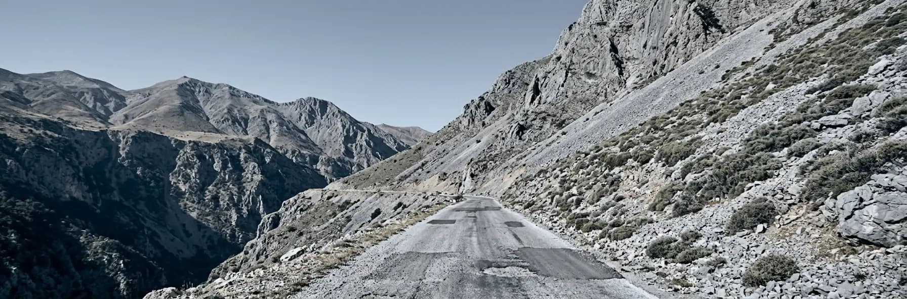 A rough patched road running along a bare grey mountainside in Albania, with a deep valley below and higher ridges beyond.