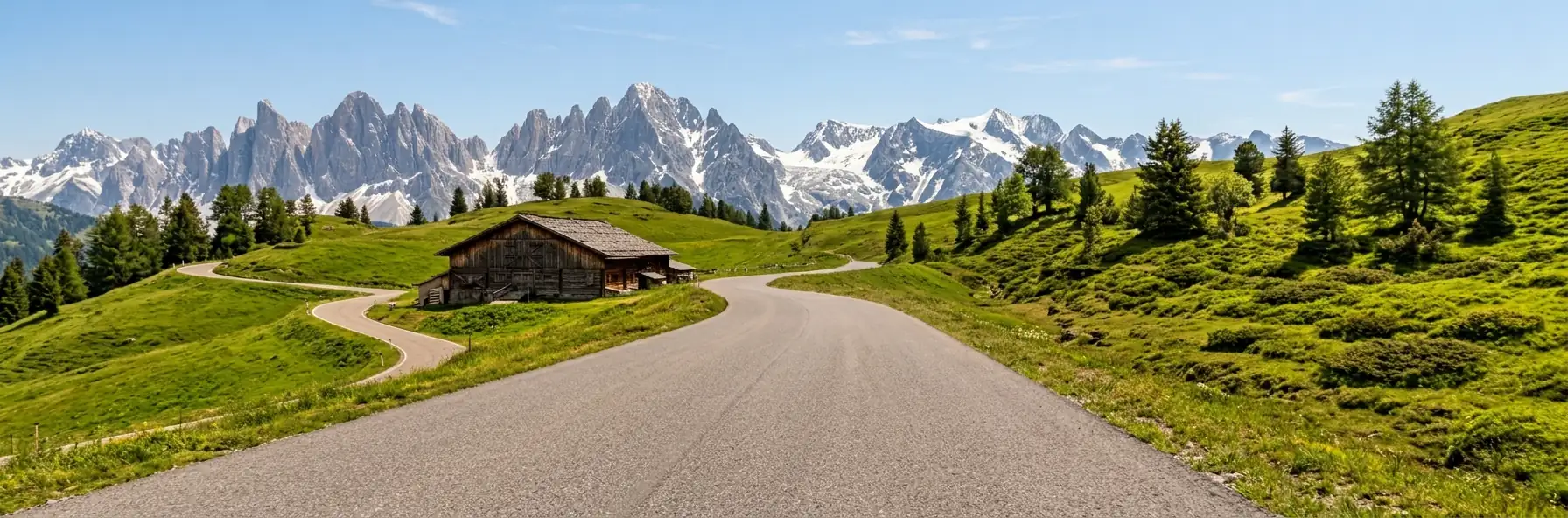 A road curving past a dark timber alpine farmhouse across green pasture in Austria, with a jagged snow-streaked mountain range on the horizon.