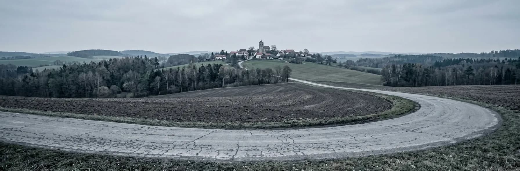 A road sweeping in a broad curve past a ploughed field in Bohemia, with a small hilltop village and church tower on the ridge beyond.
