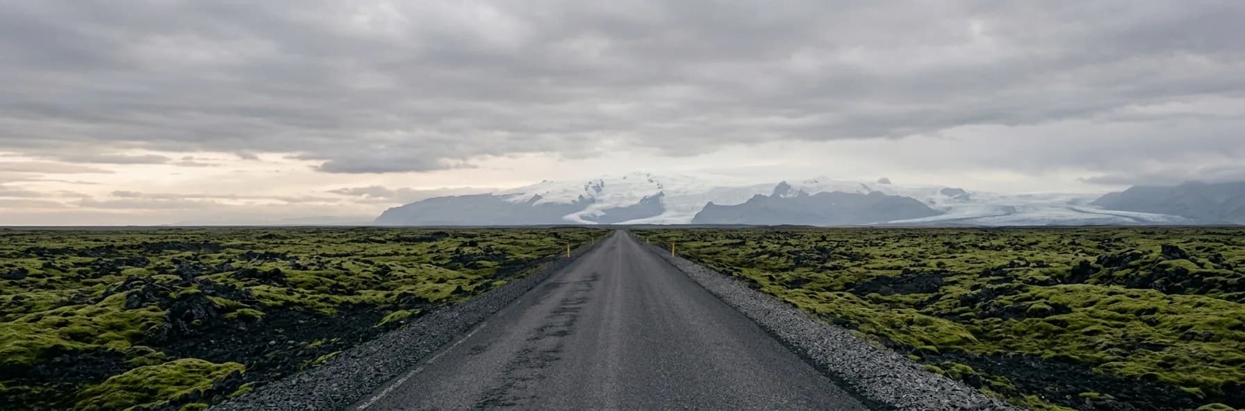 A straight road crossing a treeless volcanic plain of black lava and green moss in Iceland, with a glacier and snow-covered mountains on the horizon beneath heavy cloud.