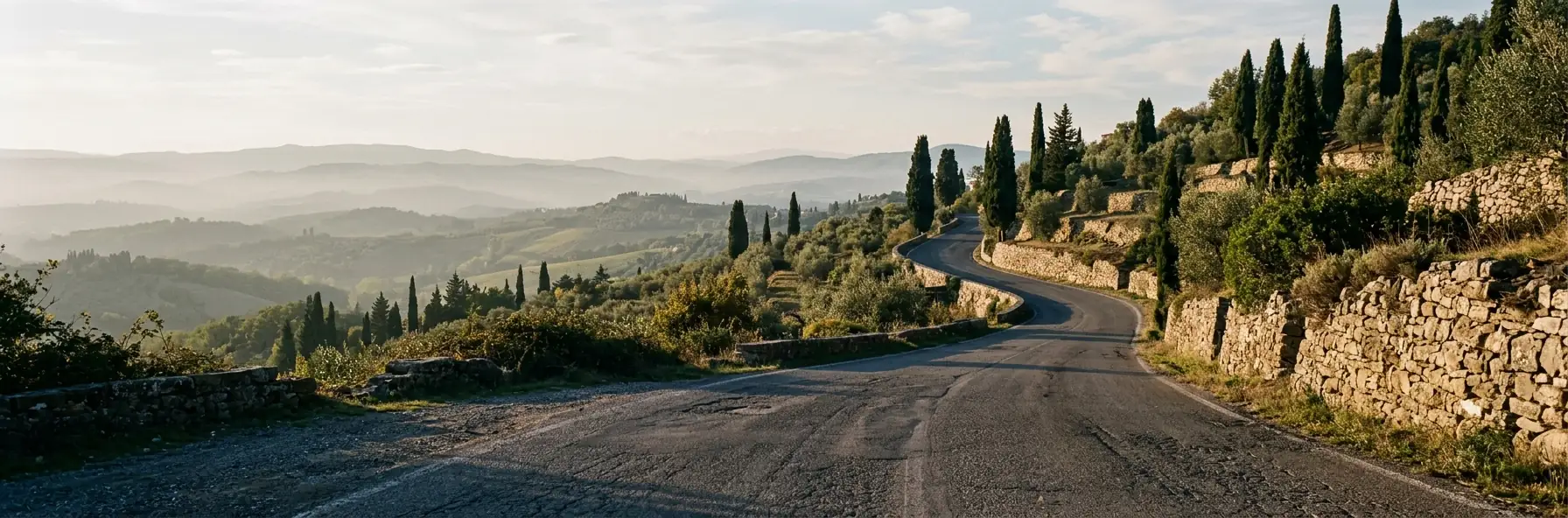 A road curving between dry-stone terraces, cypresses and olive trees on a Tuscan hillside, with hills receding into haze in the late afternoon light.