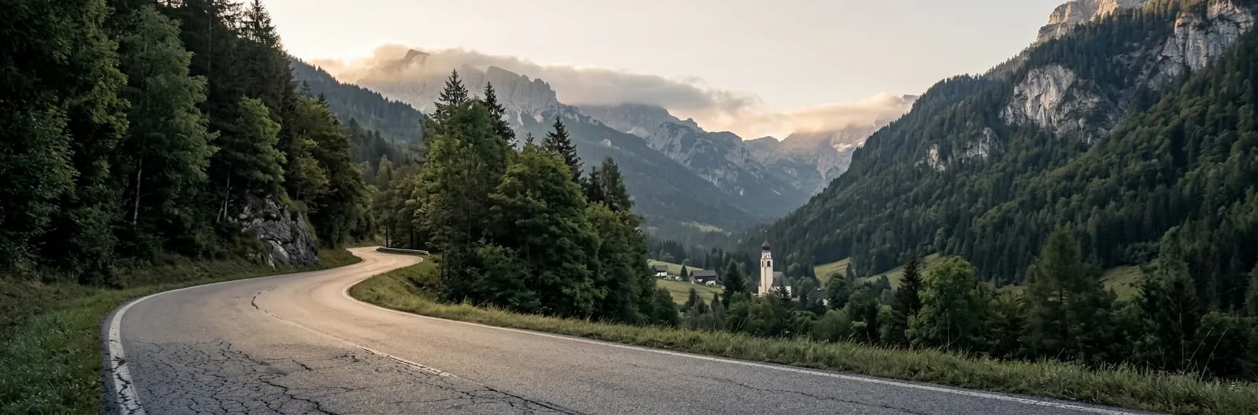 A road curving down through wooded slopes into a Slovenian alpine valley, a village church spire below and limestone peaks catching the morning light.