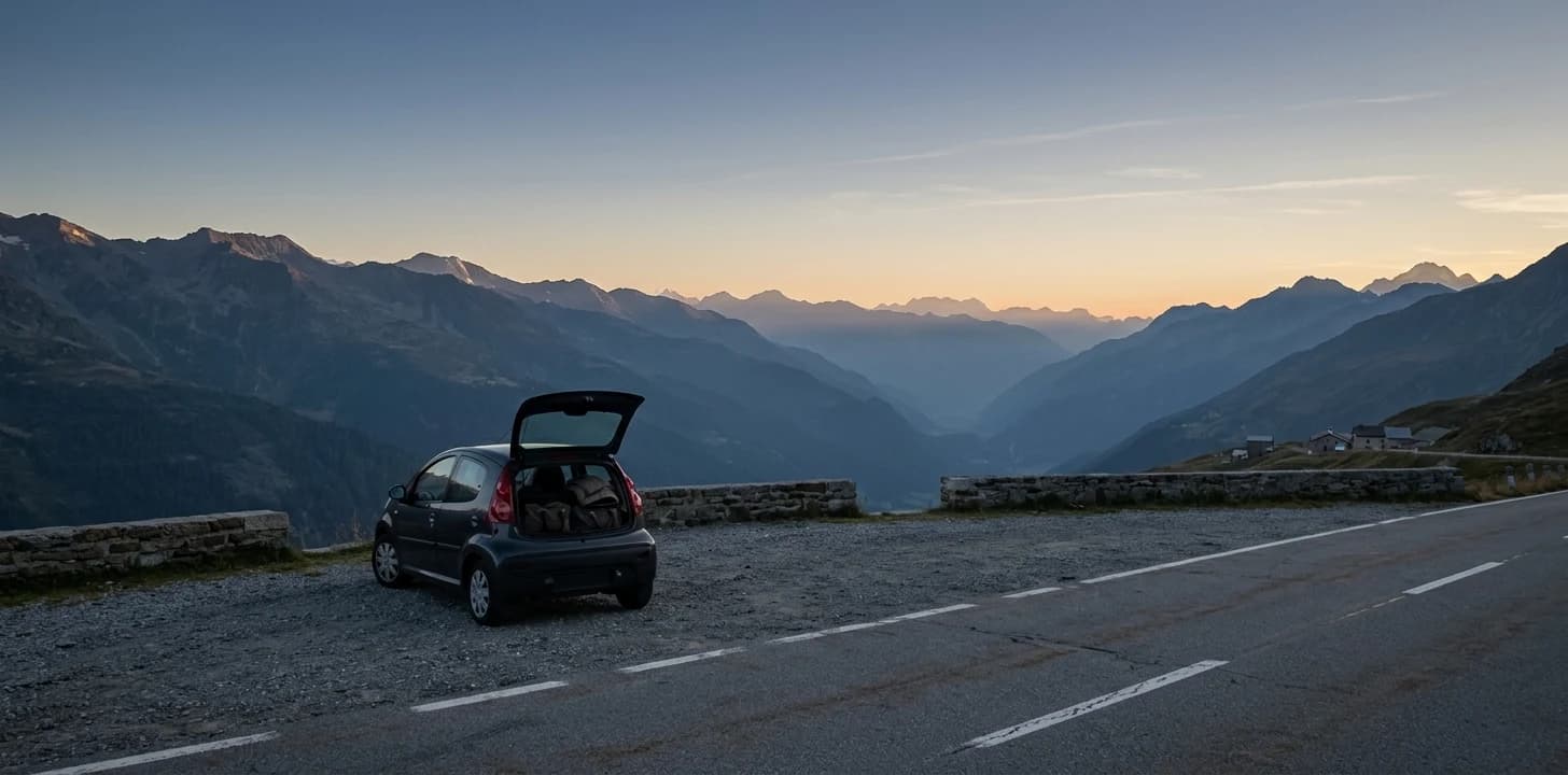 A small hatchback parked at a mountain pass viewpoint at dusk with its boot open, an alpine valley falling away below and the last light on the peaks.