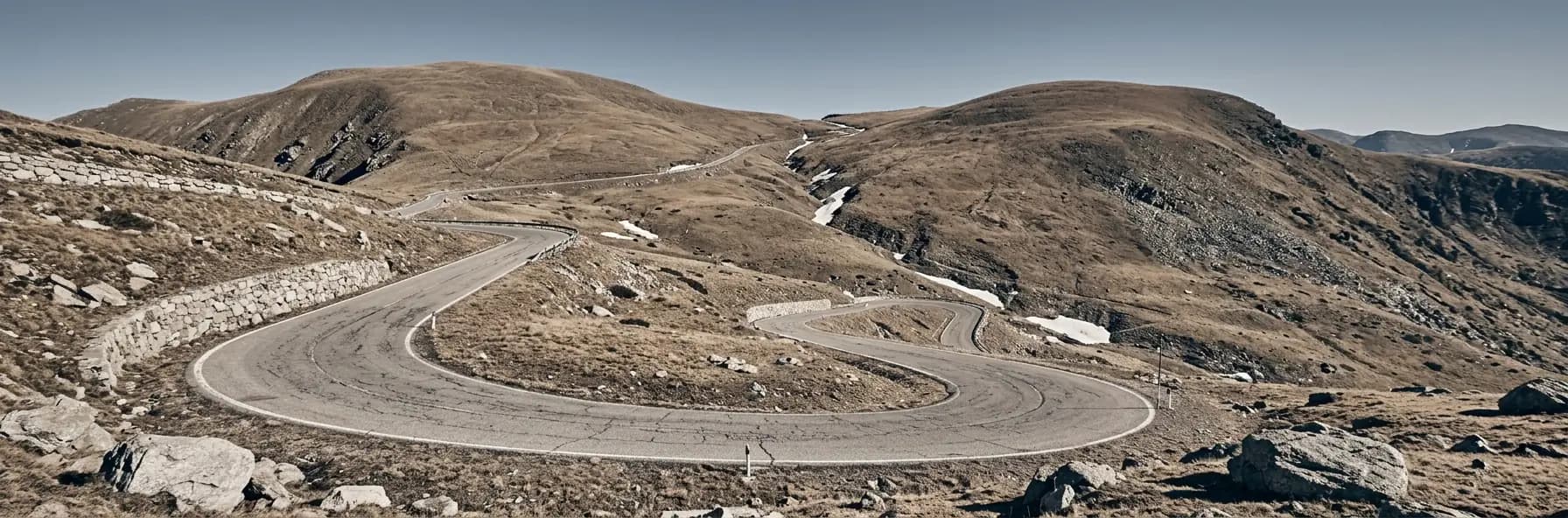 A road switchbacking across a high treeless pass in Andorra, bare tawny grass and grey rock with patches of old snow, rounded eroded Pyrenean ridges beyond.