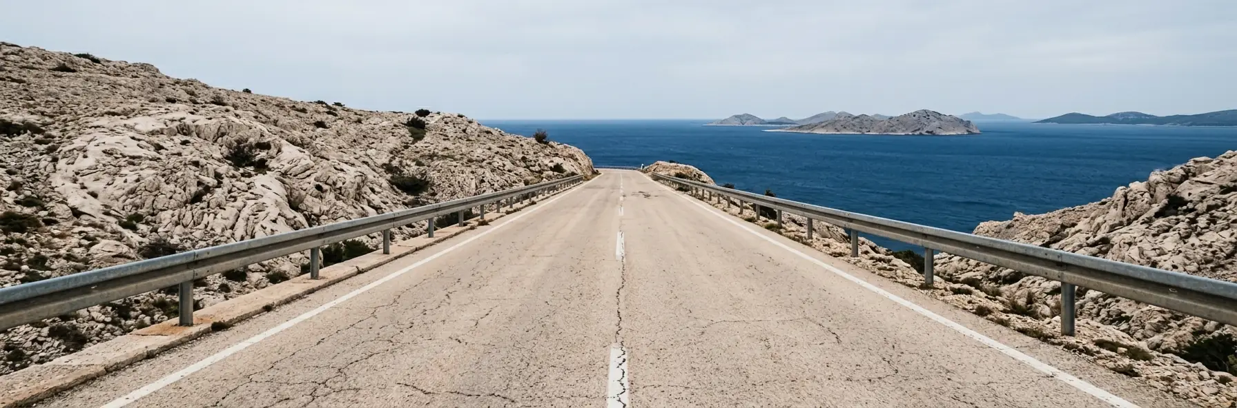 A crash-barriered road running down a bare limestone headland towards the Adriatic in Croatia, with low islands visible offshore.