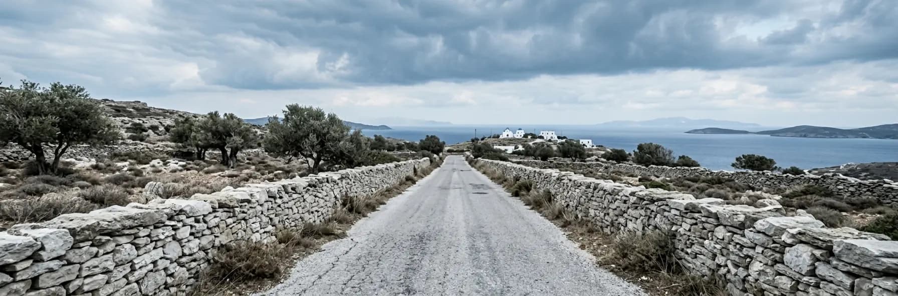 A narrow road between dry-stone walls on a rocky Aegean headland in Greece, with olive trees, whitewashed buildings and the sea beyond.