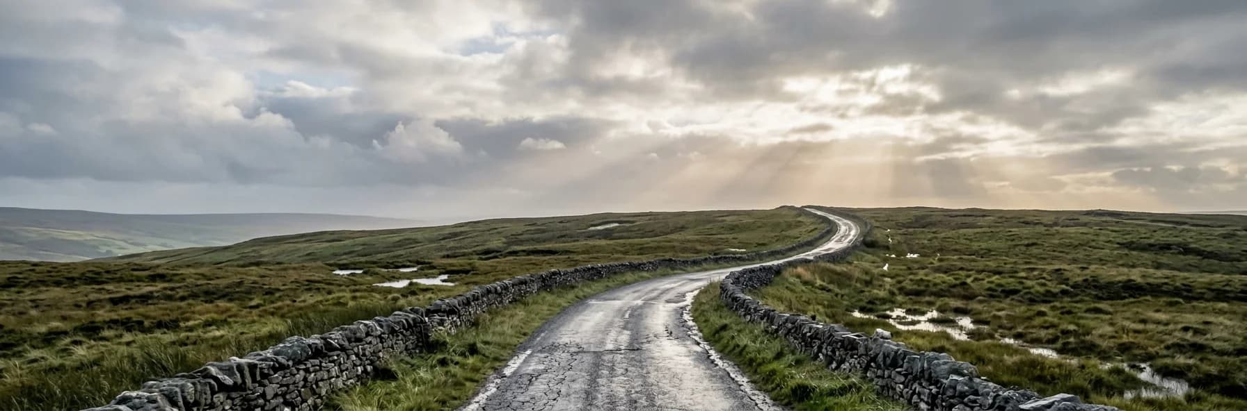 A narrow road between low stone walls curving over boggy green hillside in Ireland, wet after rain, with shafts of light breaking through cloud.