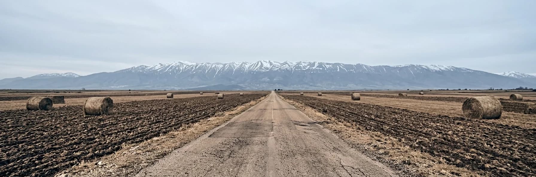 A road crossing wide open farmland in Kosovo past round hay bales, with the snow-streaked Sharr mountains along the horizon.