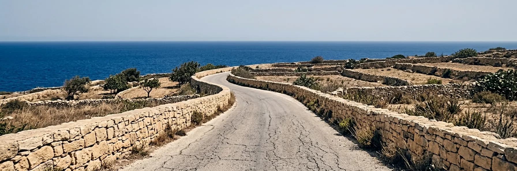 A narrow road between low honey-coloured limestone walls on Malta, small terraced fields either side and the Mediterranean beyond.