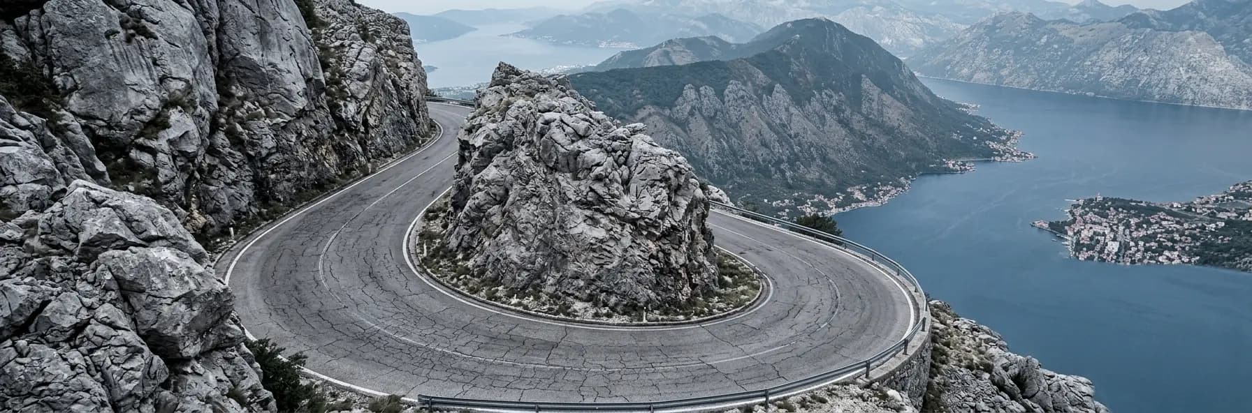 A hairpin bend on a mountain road cut into grey limestone high above the Bay of Kotor in Montenegro, the bay and a coastal town far below.