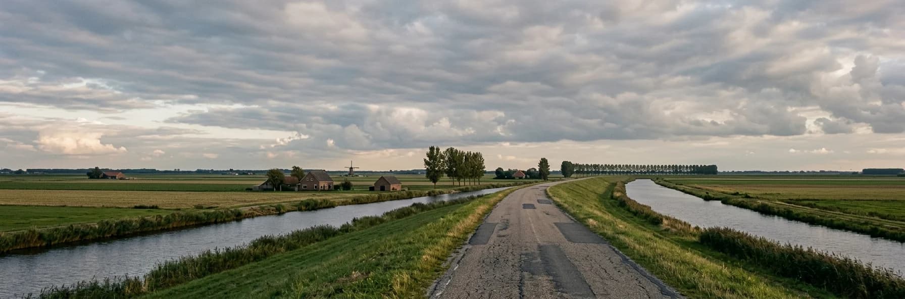 A narrow road running along the top of a grass dyke through flat Dutch polder farmland, canals on both sides and a windmill and farmhouses in the distance beneath a vast cloudy sky.