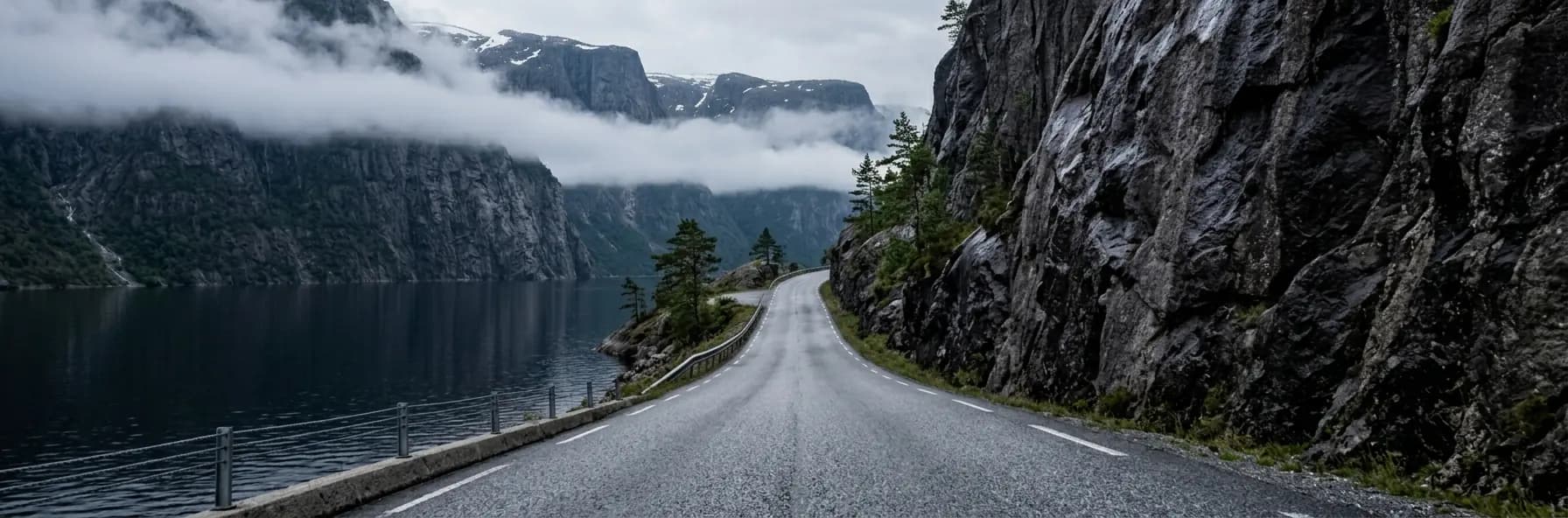 A road hugging the foot of a sheer rock face beside a deep fjord in Norway, with cloud caught halfway up the cliffs opposite.