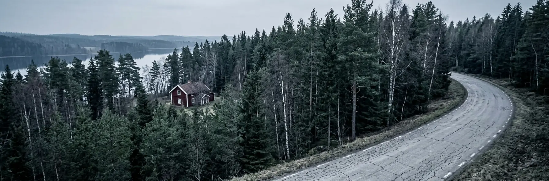 A road curving through pine and birch forest in Sweden past a red-painted timber house, with a lake visible through the trees.