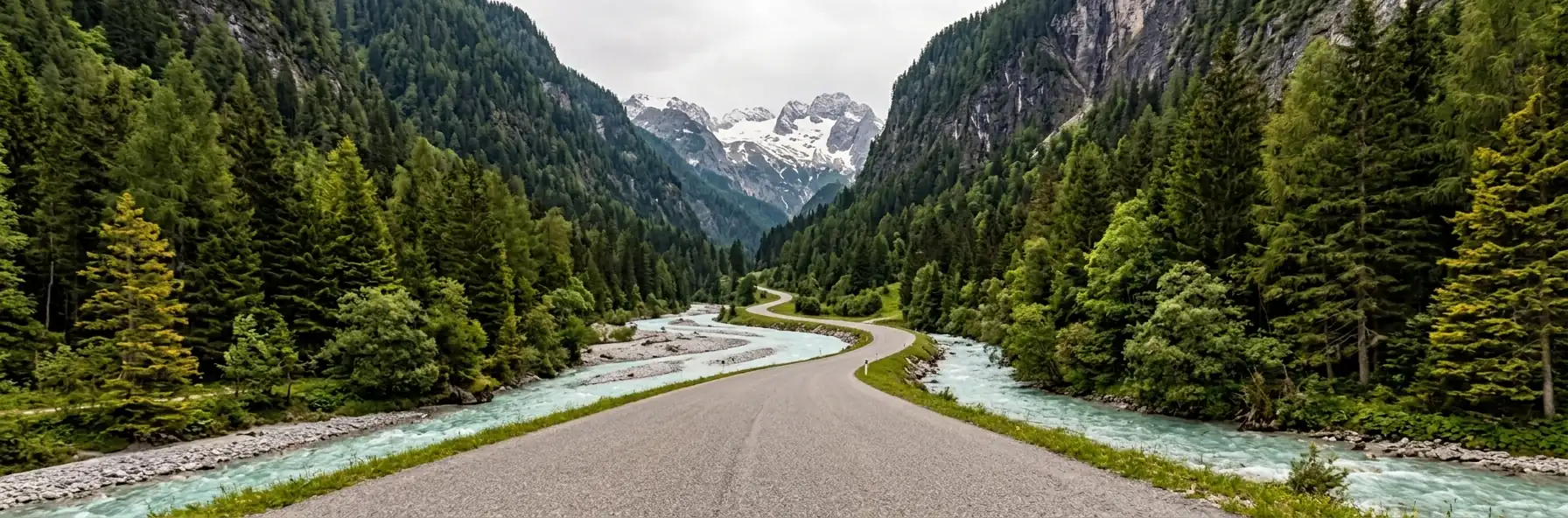 A valley road following a pale turquoise glacial river between steep forested slopes in Switzerland, with a snow-covered peak at the head of the valley.