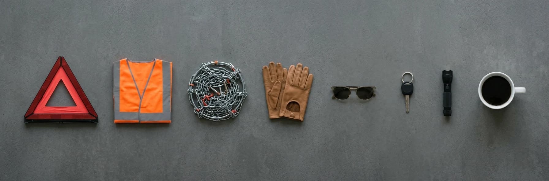 Equipment for a European road trip laid out in a row: a warning triangle, hi-vis vest, snow chains, driving gloves, sunglasses, a car key, a torch and a cup of coffee.